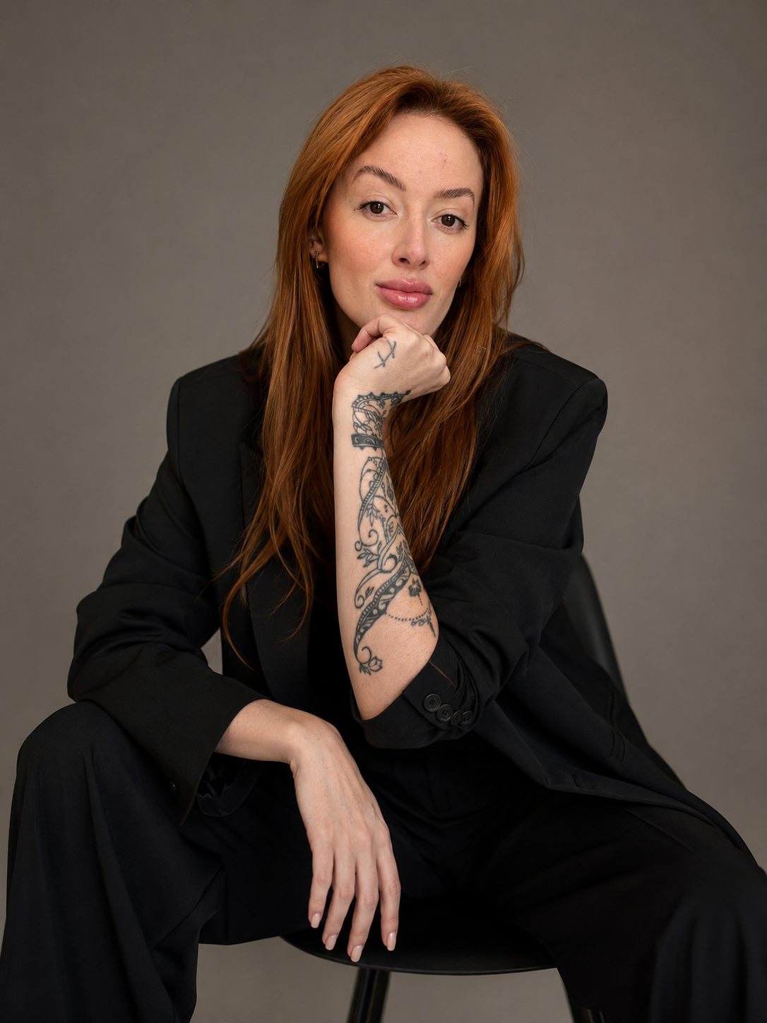 Carolina Lamarão sentada, de blazer preto, com o braço tatuado